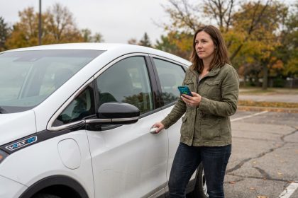 Femme évaluant une voiture électrique d'occasion dans un parking, consulte une application de recharge sur son smartphone